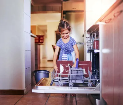 Kék-fehér ruhás mosolygó kislány tiszta tányérokat vesz ki a mosogatógépből.