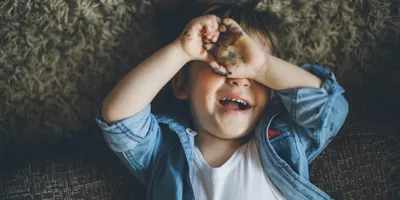 Un copil cu mâinile la ochi pătate de acuarele zâmbește. 