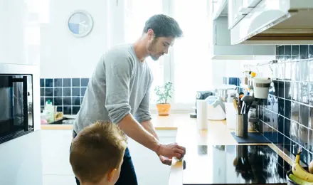 Čovjek u sivom džemperu čisti peć maramicom s djetetom pokraj sebe.