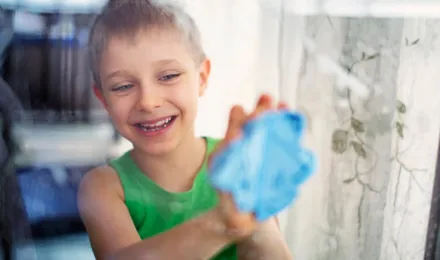 Ablaktisztítás: Mosolygó, zöld pólós kisfiú kék törlőronggyal ablakot tisztít.