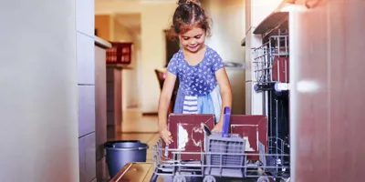 Kék-fehér ruhás mosolygó kislány tiszta tányérokat vesz ki a mosogatógépből.