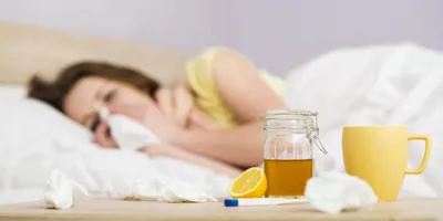 Barna hajú nő fekszik az ágyban, az orrát fújja, előtte az asztalon a megfázás kezelésére szolgáló méz és tea.