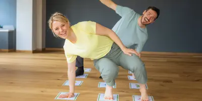Lachende Frau und Mann machen spielerische Yoga-Posen auf illustrierten Karten auf einem Holzboden.