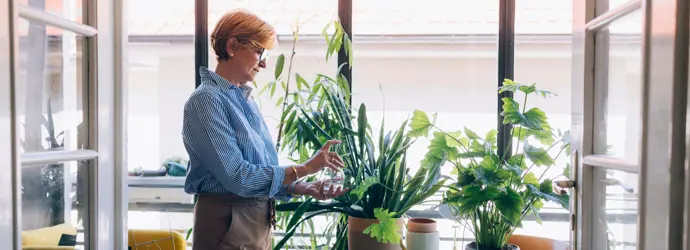 Eine Frau mit kurzen roten Haaren und Brille besprüht Pflanzen, die auf einem weißen runden Tisch in einem Wohnraum stehen.