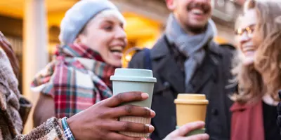 Lachende Freunde halten draußen Kaffeetassen.