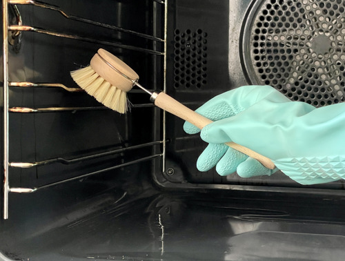 Eine mit einem türkiesen Haushaltshandschuh geschützte Hand schrubbt die Seitenwand eines Ofens mit einer hölzerne Spülbürste.