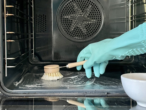 Eine mit einem türkiesen Haushaltshandschuh geschützte Hand schrubbt den Boden des Ofens mit einer Holzbürste.