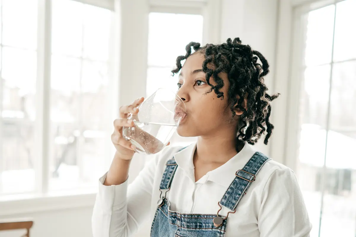 Mladá osoba stojí v jasně osvětlené místnosti a pije sklenici vody.