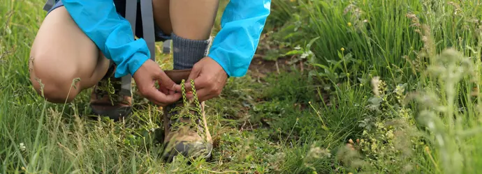 Eine Person in einem leuchtend blauen Regenmantel kniet auf dem Gras und bindet ihre grün-schwarzen Schnürsenkel.