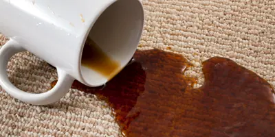 Eine umgekippte weiße Tasse vergießt ihren Inhalt auf einen beigen Teppich.