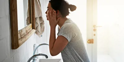 Szürke felsőben lévő nő mossa az arcát egy fehér mosdókagyló felett.