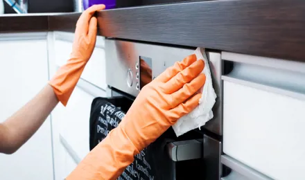 Mâini cu mănuși din cauciuc se sprijină de partea de sus a unui cuptor și cu o cârpă curăță cuptorul. 
