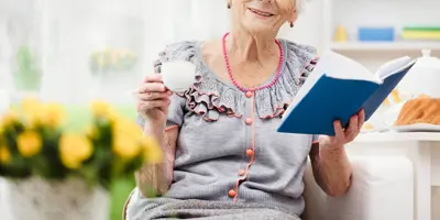 O femeie în vârstă, zâmbitoare, ține o ceașcă de ceai într-o mână și o carte deschisă în cealaltă, într-un decor luminos și liniștit.