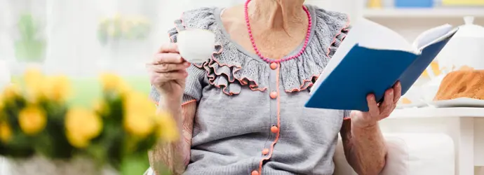 O femeie în vârstă, zâmbitoare, ține o ceașcă de ceai într-o mână și o carte deschisă în cealaltă, într-un decor luminos și liniștit.