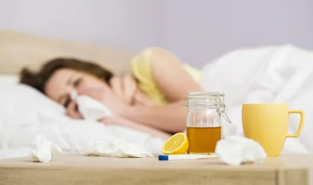 Femeie răcită în pat suflându-și nasul. O cană, un borcan cu miere și lămâie pe o masă alăturată.