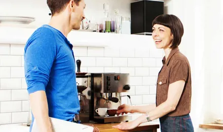 Μια γυναίκα χρησιμοποιεί μια καφετιέρα, ενώ μιλάει με έναν άντρα στην κουζίνα