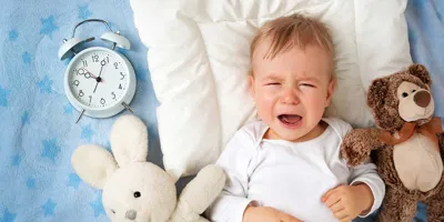 Ένα μωρό πάνω σε μια κουβέρτα κλαίει ενώ περιβάλλεται από δύο λούτρινα παιχνίδια και ένα αναλογικό ξυπνητήρι
