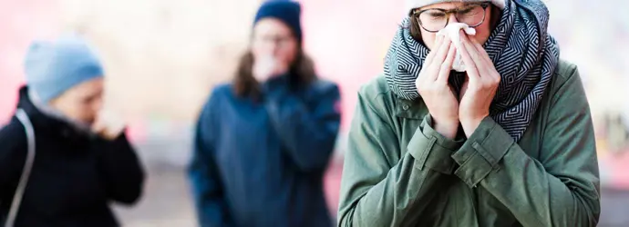 Három, az utcán sétáló ember látszik, kettő köhög, az elöl álló személy az orrát fújja.