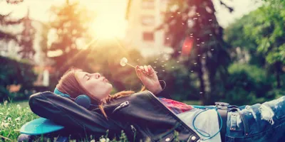 Молодая женщина, лежа на траве, слушает музыку и дует на одуванчик