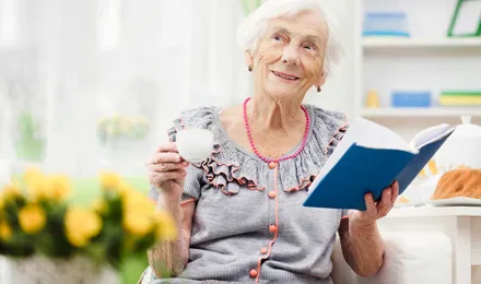 Пожилая женщина сидит на диване, держа книгу и чашку