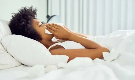 Crnkinja leži u krevetu s bijelom plahtom i drži maramicu na nosu.