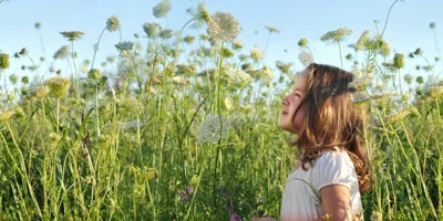 O tânără fată care ar putea avea nevoie de remedii pentru alergie la polen, aflându-se pe un câmp plin de flori inalte, sălbatice, într-o zi însorită