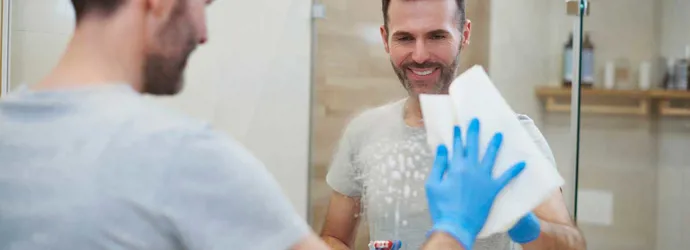 Женщина в синих перчатках чистит зеркало в ванной комнате