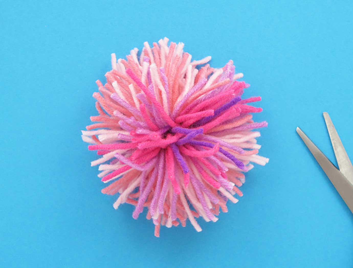 Gotov pompom, napravljen od različiih nijansa ružičaste vune, uz škare sa strane.
