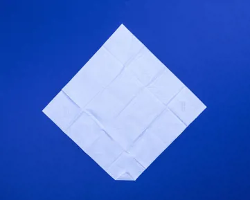 Fehér zsebkendő kiterítve, átlós helyzetben, az alsó sarka behajtva, kék asztalon.