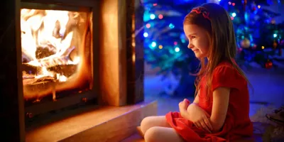 Mosolygó, piros ruhás kislány a megrakott kandalló előtt ül, mögötte karácsonyfa látszódik.