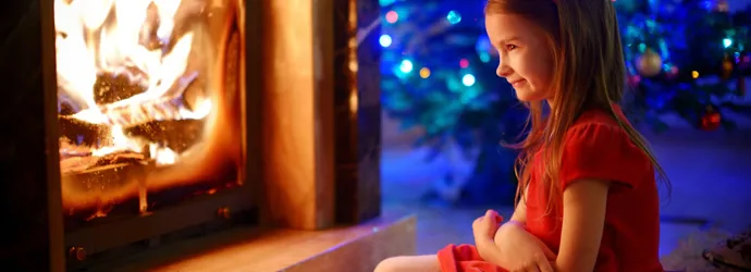 Mosolygó, piros ruhás kislány a megrakott kandalló előtt ül, mögötte karácsonyfa látszódik.