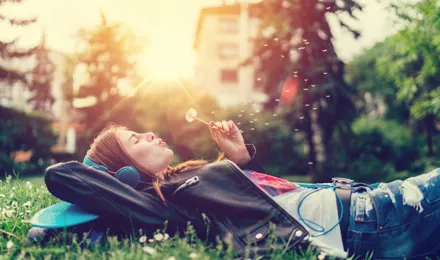 A menstruációs görcsök enyhítése relaxálással: Fiatal nő a fűben fekve zenét hallgat, és elfúj egy pitypangot.