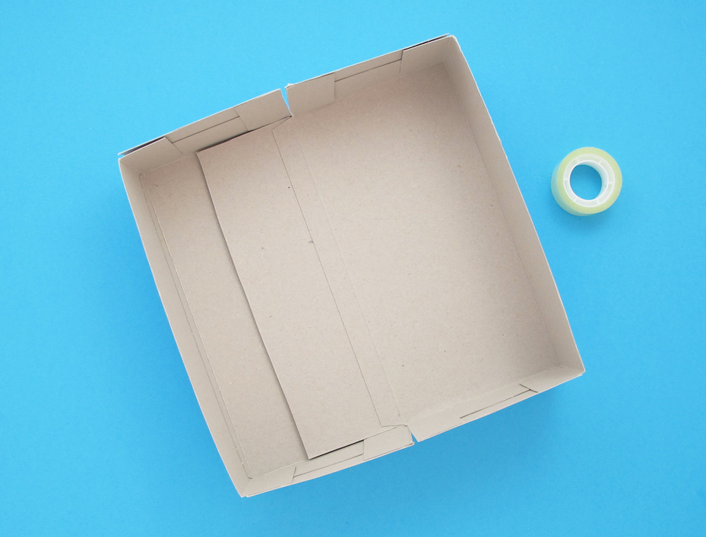 Egy ragasztószalaggal megerősített kartondoboz egy átlátszó ragasztószalag-tekercs mellett, kék háttér előtt.