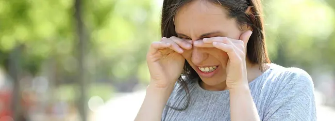 Szürke pólóban lévő barna hajú nő a kézfejével dörzsöli viszkető, piros szemeit.