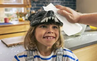 Ein Kind trägt eine Tiermaske auf dem Kopf, während ein Mann ein Blatt Küchenpapier benutzt, um etwas davon abzuwischen.