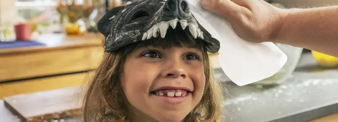 Ein Kind trägt eine Tiermaske auf dem Kopf, während ein Mann ein Blatt Küchenpapier benutzt, um etwas davon abzuwischen.