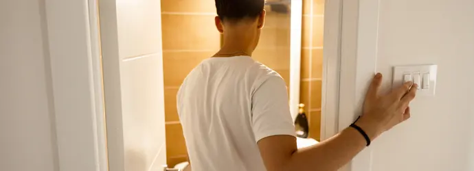 Ein Mann in einem weißen T-Shirt betritt ein Badezimmer und schaltet gleichzeitig das Licht ein.