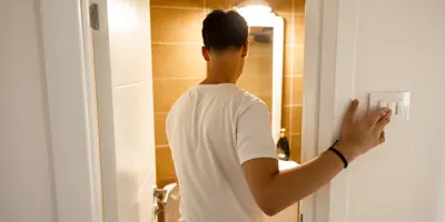 Ein Mann in einem weißen T-Shirt betritt ein Badezimmer und schaltet gleichzeitig das Licht ein.