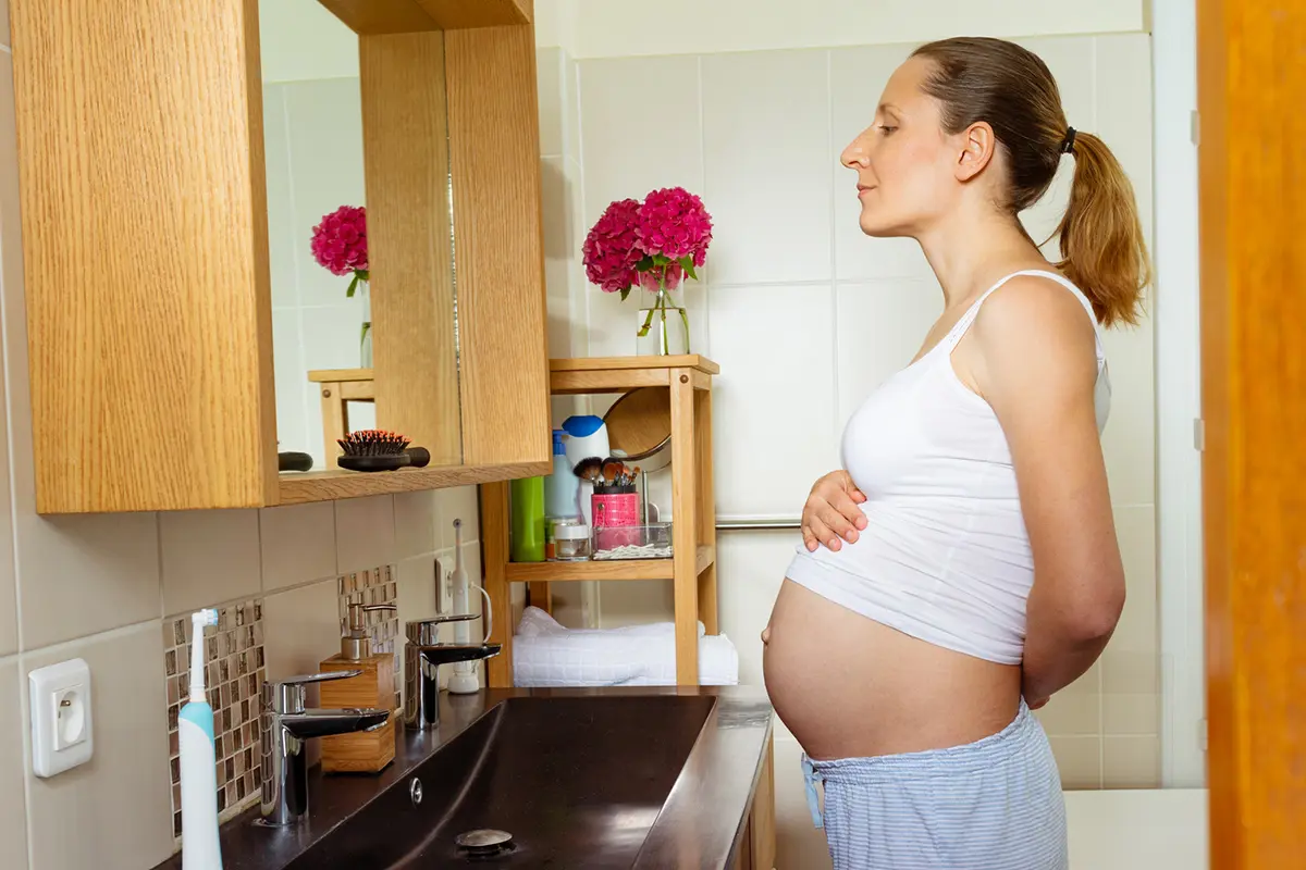 Těhotná žena stojí před zrcadlem, horní díl má zvednutý přes těhotenský břich.
