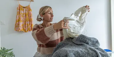 Eine blonde Frau in Wollpullover und Jeans sitzt in einem Zimmer und hält ein weißes Kleidungsstück in den Händen.