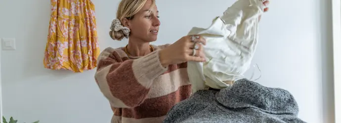 Eine blonde Frau in Wollpullover und Jeans sitzt in einem Zimmer und hält ein weißes Kleidungsstück in den Händen.