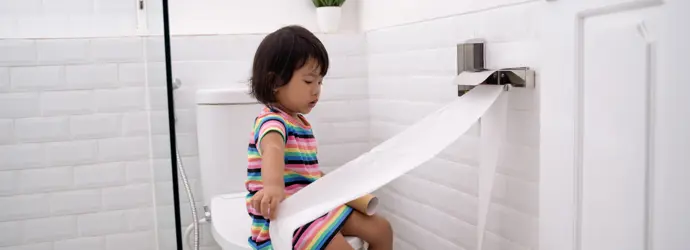 Egy kislány WC-papírt húz ki a WC-n ülve.