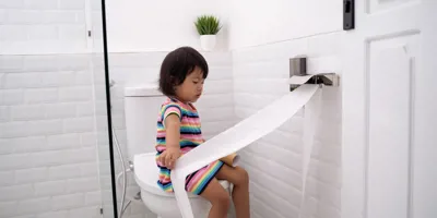 Egy kislány WC-papírt húz ki a WC-n ülve.
