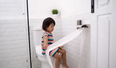 Egy kislány WC-papírt húz ki a WC-n ülve.