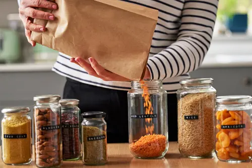 Μια γυναίκα ρίχνει φακές από μια χάρτινη σακούλα σε ένα βάζο, δίπλα σε άλλα βάζα