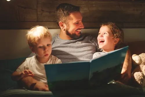 Ein Mann liest zwei kleinen Kindern im Bett vor, eines neben ihm. Ein Kind und der Mann lächeln.