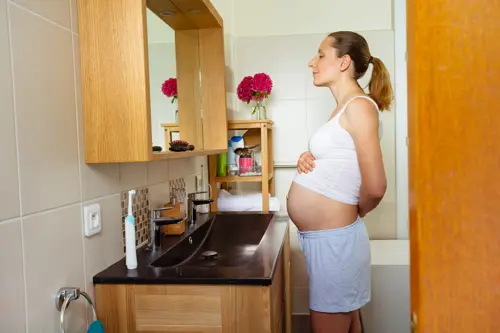 Eine schwangere Frau steht vor einem Spiegel und hat ihr Oberteil über ihren Schwangerschaftsbauch gezogen.
