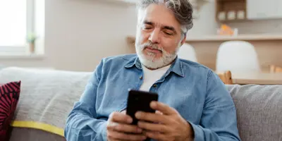 Ein älterer Mann in einem Jeanshemd sitzt lächelnd auf einer Couch und hält ein Handy in der Hand.
