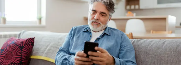 Ein älterer Mann in einem Jeanshemd sitzt lächelnd auf einer Couch und hält ein Handy in der Hand.