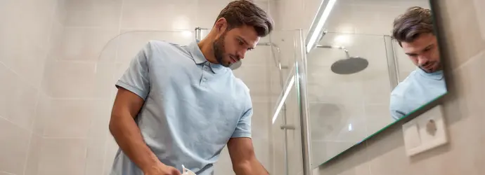 Ein Mann in einem Poloshirt und Jeans reinigt eine Badezimmerarmatur mit einer Sprühflasche und einem Lappen.